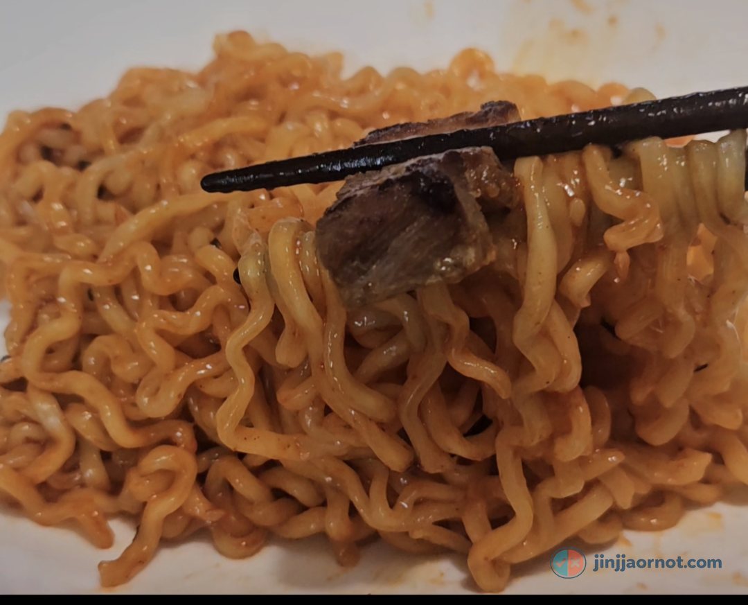 Chopsticks lifting a piece of grilled pork belly over a plate of Original Buldak noodles in red sauce