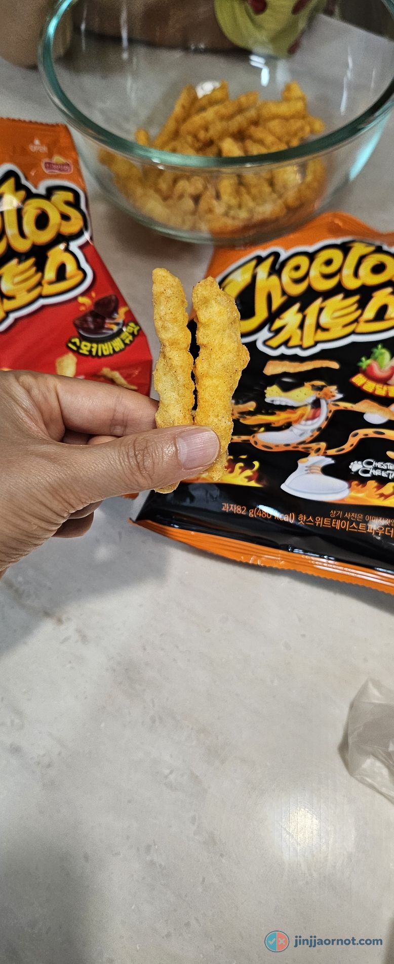A hand holding two Korean Cheetos sticks up close, showing the pale golden seasoning rather than bright orange cheese dust