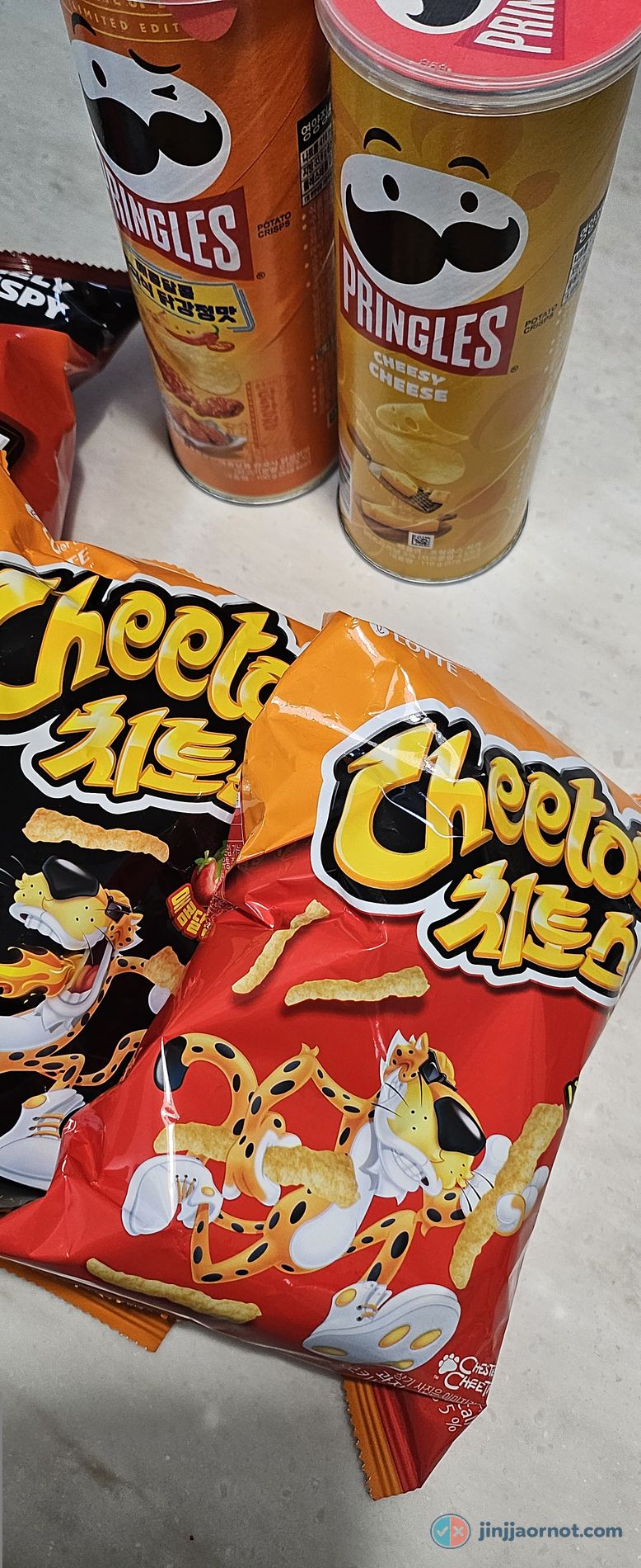 Two bags of Korean Lotte Cheetos on a counter — Smoky BBQ in a red bag and Spicy Sweet in a black bag — beside a glass bowl filled with the puffed corn sticks