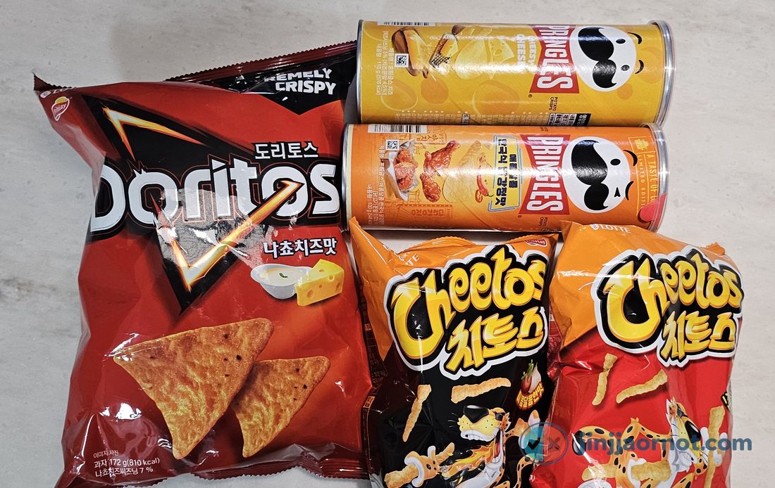 Korean versions of American snacks laid out together: a bag of Doritos Nacho Cheese, two cans of Pringles including a Korean-style dakgangjeong limited edition, and two bags of Lotte Cheetos