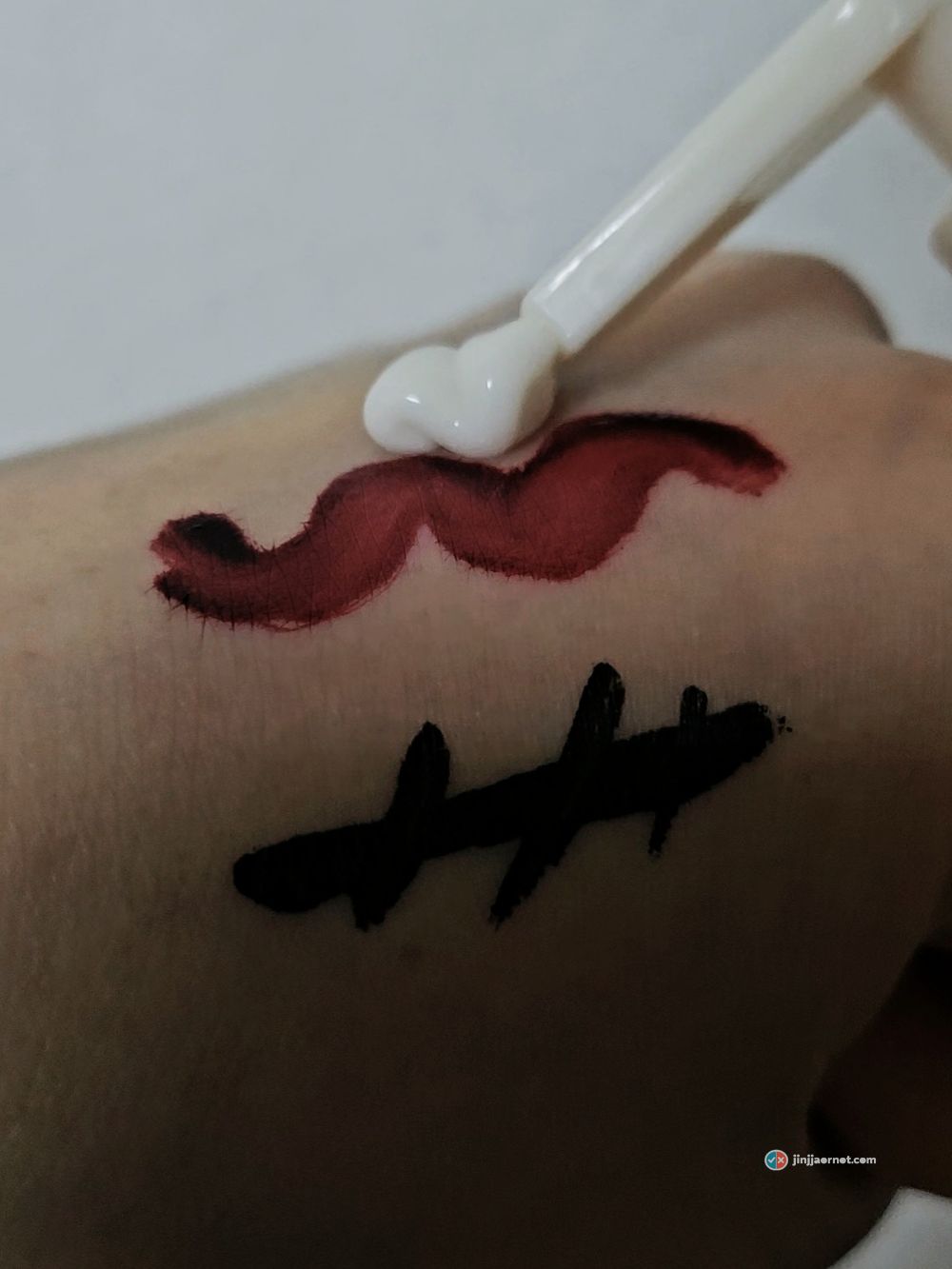 Cleansing milk placed with a spatula onto a wavy stripe of long-wear lip tint drawn on the back of a hand