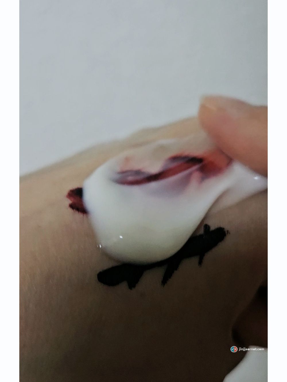 Thick white cleansing milk sitting on top of lip tint on the back of a hand, colour bleeding up into it