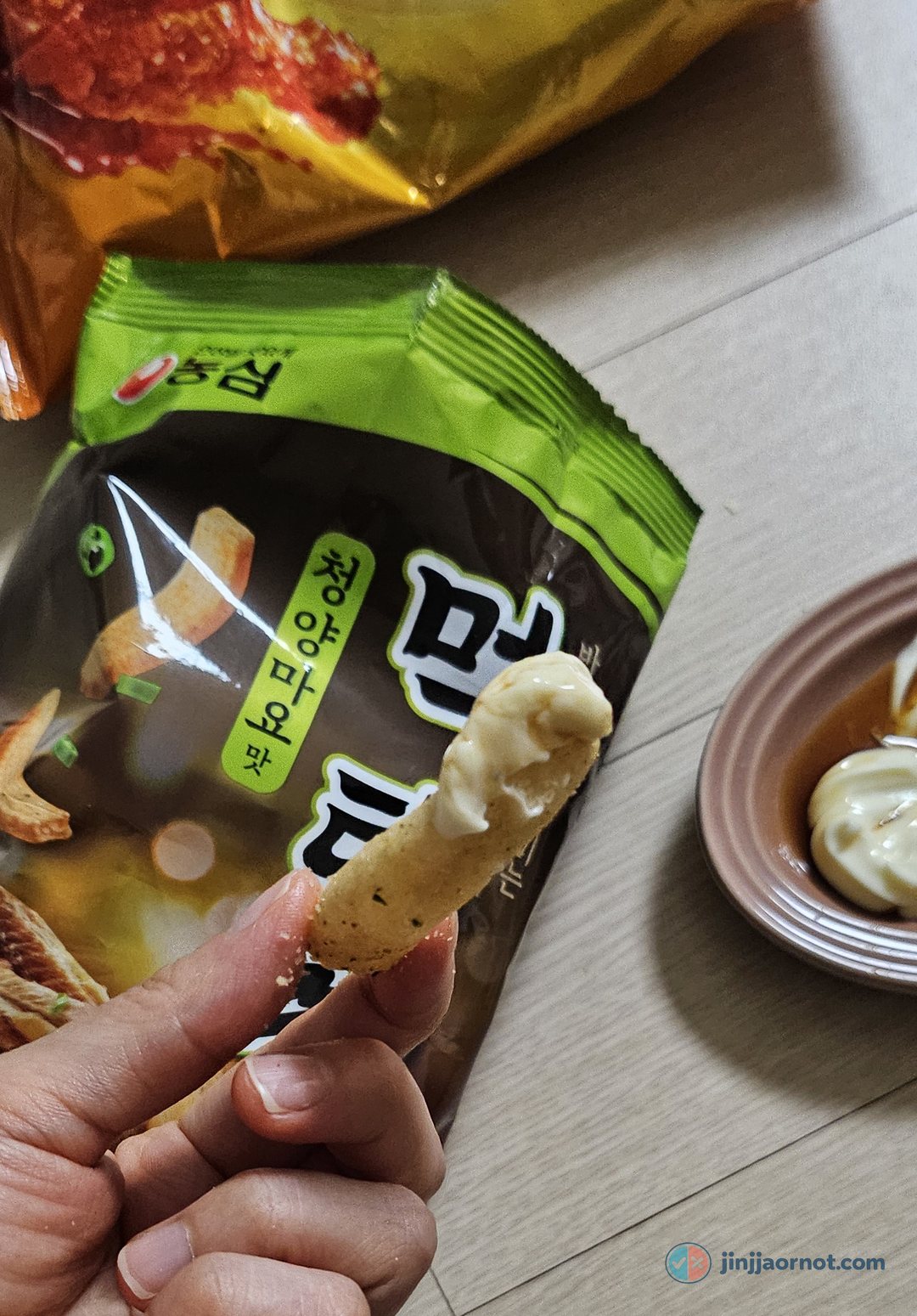 A hand holding a Mektaekkang chip dipped in white mayonnaise, with the bag and a plate of mayo in the background