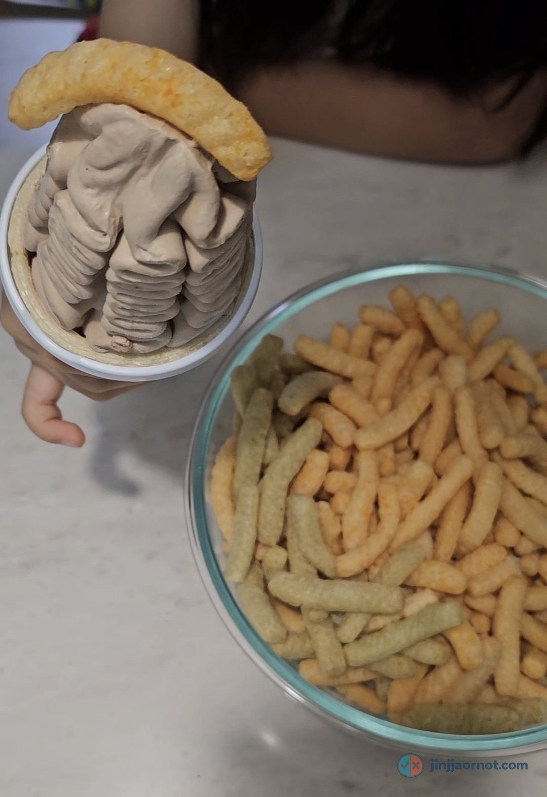 A hand holding a cup of chocolate soft-serve with a shrimp chip resting on top, beside a glass bowl of mixed Saewookkang chips