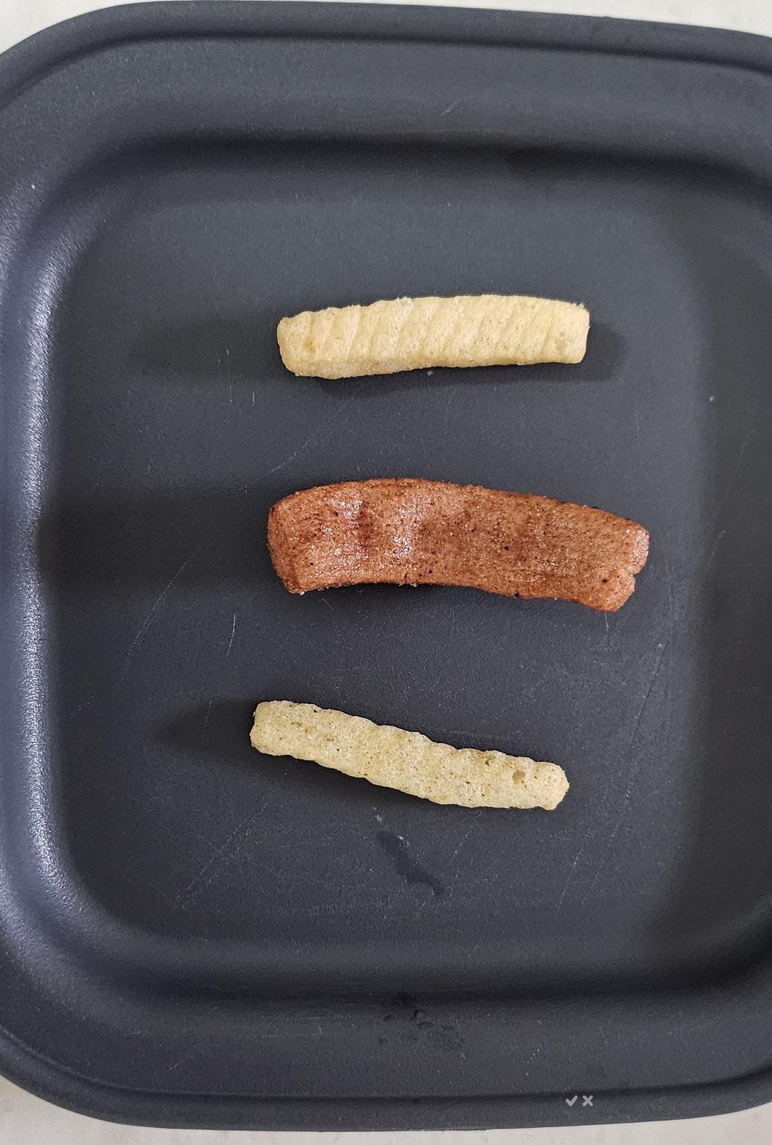 Three different Nongshim chips laid on a dark tray: a pale thin Rice Saewookkang stick, a wide reddish-brown Yukpokkang jerky piece, and a green Wasabi Saewookkang stick